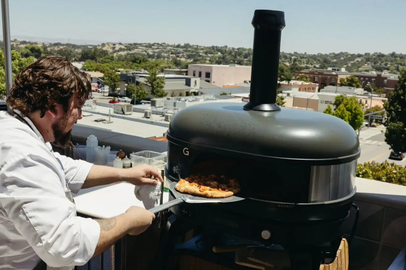 A chef uses a pizza peel to remove a baked pizza from a modern outdoor pizza oven on a rooftop, with a scenic view of buildings and trees in the background.