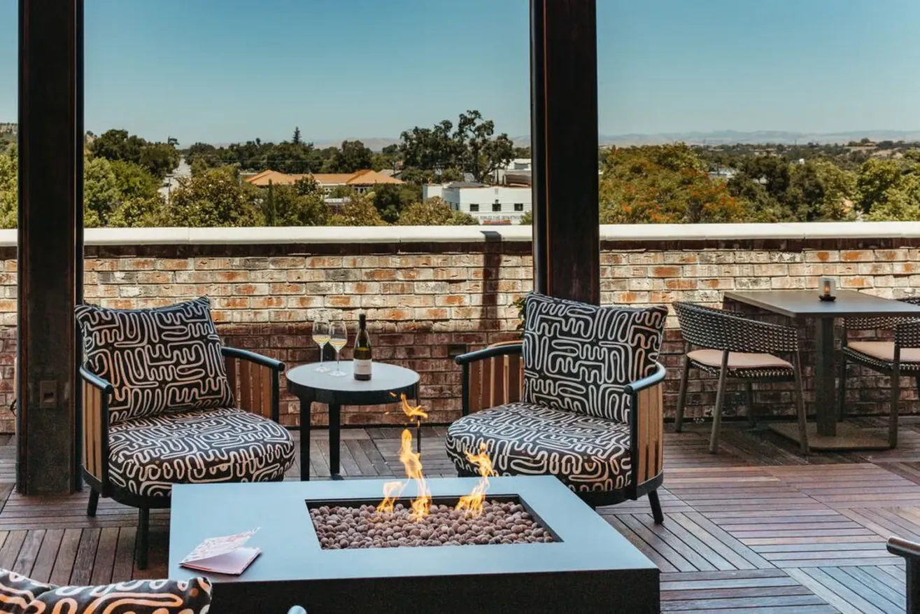 Two patterned armchairs sit by a square fire pit on a wooden rooftop patio, with a small table holding wine glasses and a bottle—perfect for relaxing after exploring Paso Robles restaurants, against views of trees and distant hills.