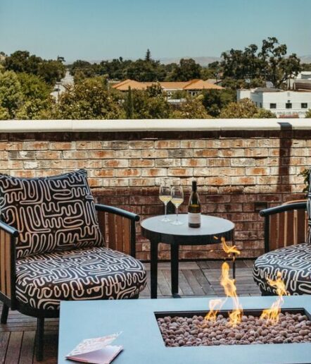Two patterned armchairs and a small table with wine glasses and a bottle sit on a patio with a fire pit. A brick wall encloses the space, and trees and rooftops are visible in the background.