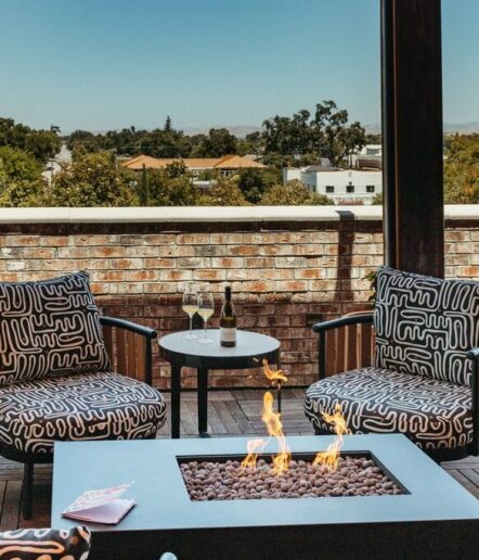 Two patterned armchairs and a small table with wine glasses and a bottle are arranged on a patio with a brick wall and a fire pit, overlooking trees and buildings under a clear sky.