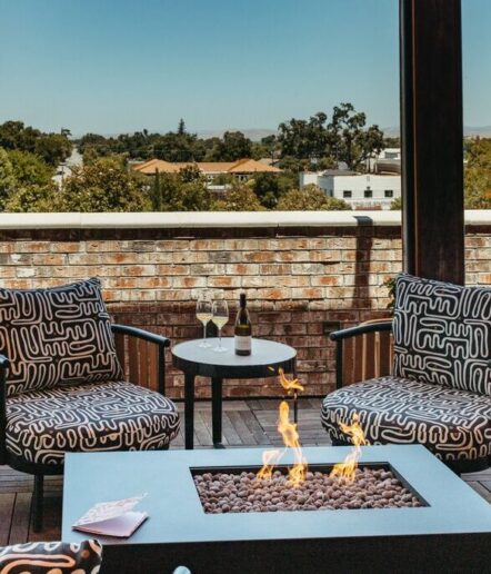 Two modern patterned armchairs surround a square fire pit on a patio, with a small table between them holding two wine glasses and a bottle. A brick wall and trees are in the background under a clear sky.