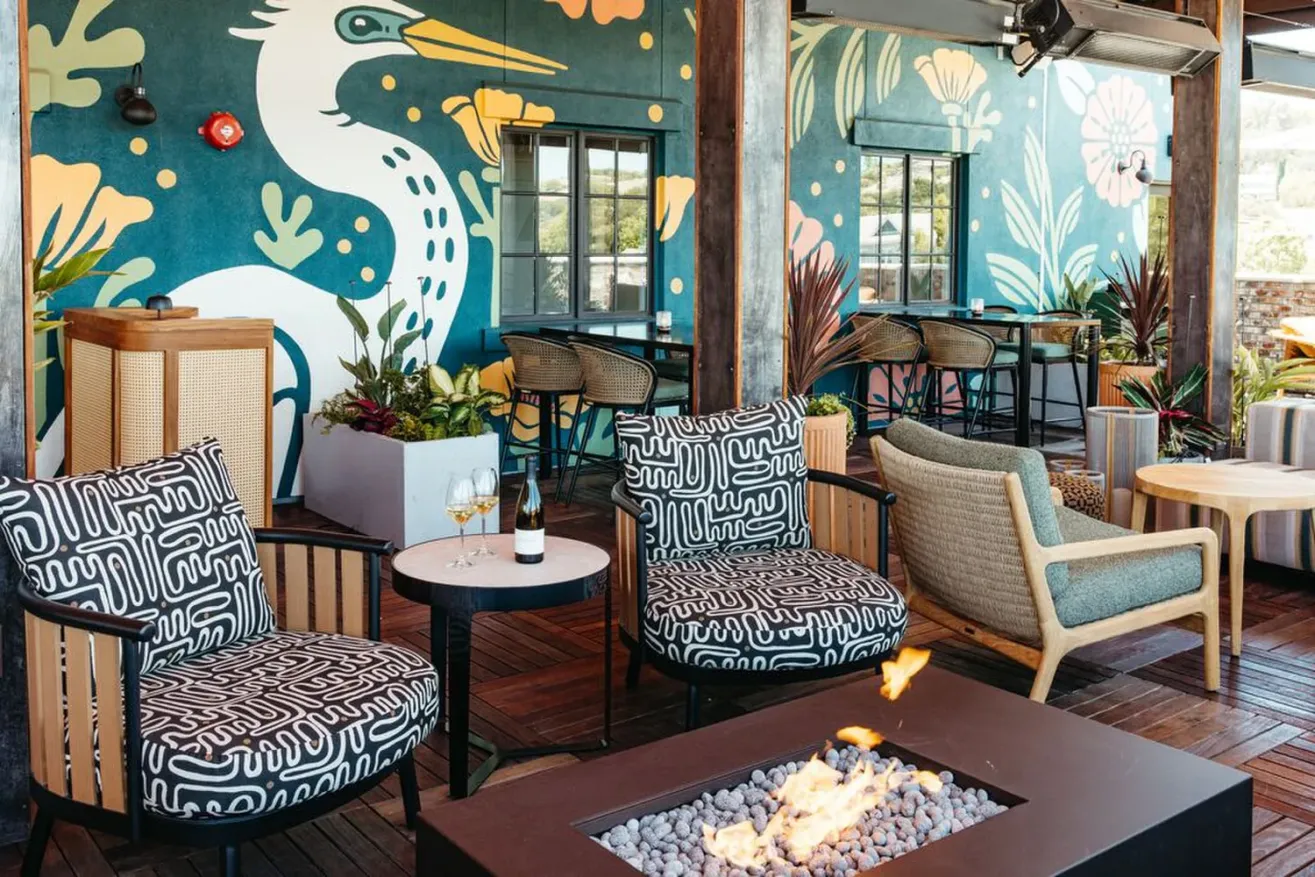 Colorful outdoor lounge area with patterned chairs, a round table with drinks, a fire pit, and a vibrant mural of a bird and flowers on the wall in the background.