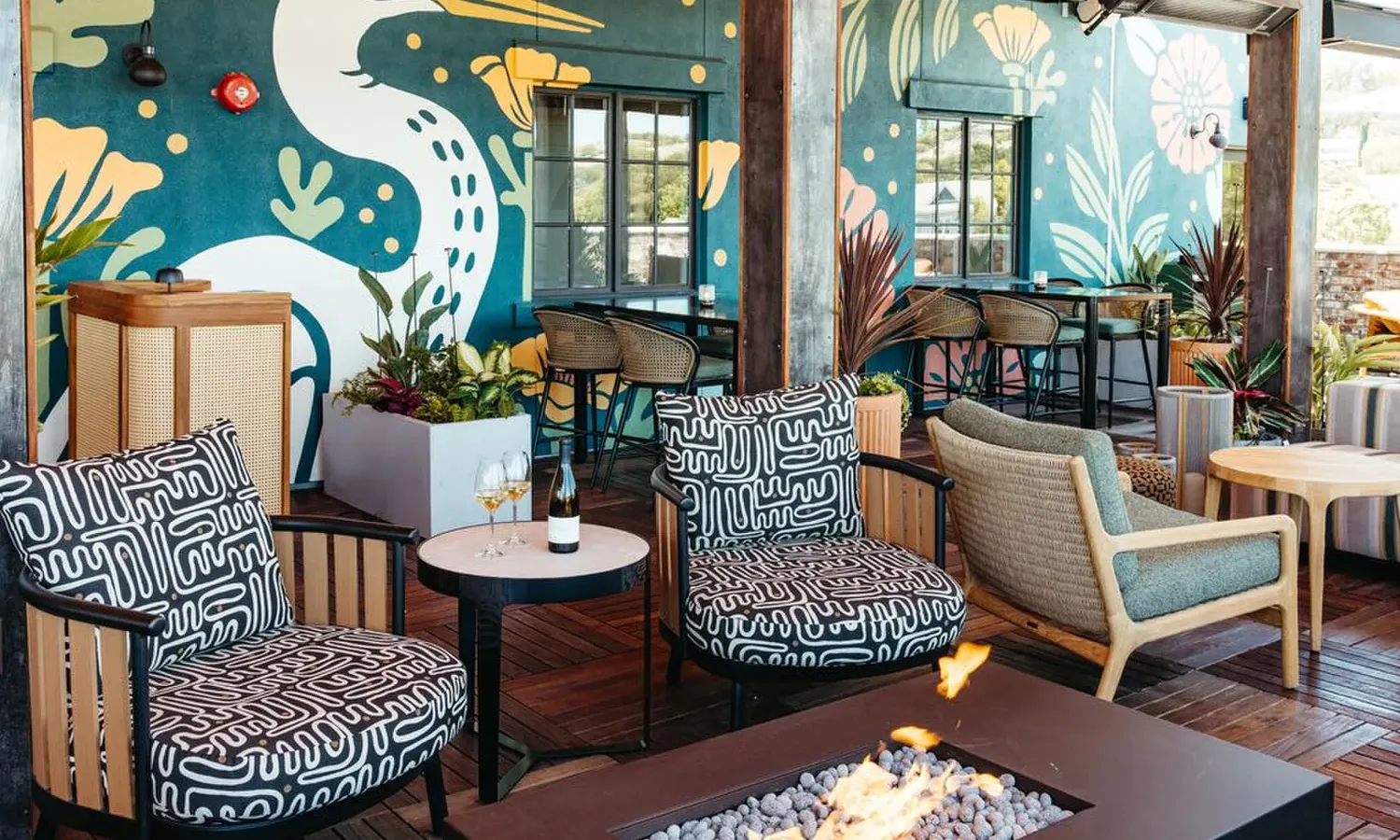 Colorful outdoor lounge area with patterned chairs, a round table with drinks, a fire pit, and a vibrant mural of a bird and flowers on the wall in the background.