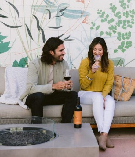 A man and woman sit on an outdoor couch, smiling and drinking wine. The woman wears a yellow sweater and white pants, and the man wears a beige jacket. A wine bottle sits on a table in front of them. A plant mural decorates the wall.