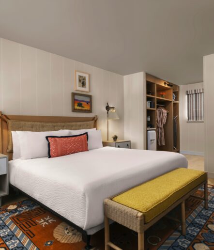 A neatly made bed with white linens, a red accent pillow, and a cane headboard stands in a cozy bedroom. The room includes a yellow bench, colorful rug, wall art, nightstands, and an open closet with robes and a mini fridge.