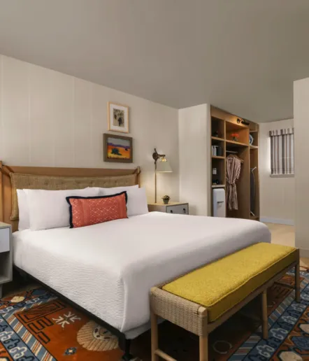 A neatly made bed with white linens, a red accent pillow, and a cane headboard stands in a cozy bedroom. The room includes a yellow bench, colorful rug, wall art, nightstands, and an open closet with robes and a mini fridge.