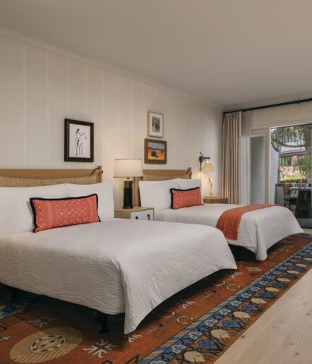 A cozy hotel room with two double beds, white bedding, orange accent pillows and throws, a patterned rug, bedside tables with lamps, wall art above the beds, and sliding glass doors leading to an outdoor patio.