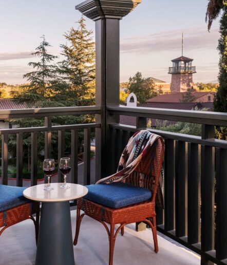 Two chairs with blue cushions and a small table holding two glasses of red wine sit on a balcony overlooking trees and a distant building with a tower at sunset.