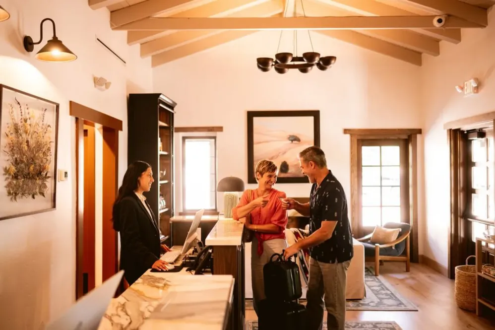 A couple with luggage checks in at a hotel reception desk while a receptionist assists them—just one example of the welcoming experiences found through Careers with Peregrine Hospitality. The room is warmly lit with wooden beams, wall art, and large windows letting in natural light.