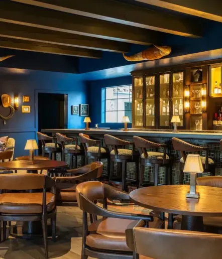 A stylish bar with wooden tables and chairs, cushioned barstools, dim lighting, wall art, and a cabinet displaying bottles. Blue walls and ceiling beams add a modern, cozy atmosphere.