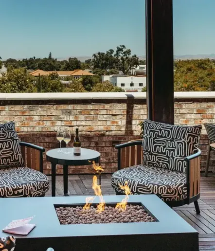 Two patterned lounge chairs sit beside a small table with wine and glasses on a patio. A fire pit burns in the foreground, and trees and rooftops are visible beyond a brick wall.
